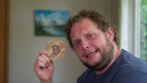 Foto van presentator, je ziet alleen zijn hoofd, die een Bitcoin vasthoudt.