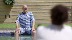 Foto van een man die zit aan de rand van een zwembad, je ziet nog de rug van iemand, maar niet scherp in beeld.