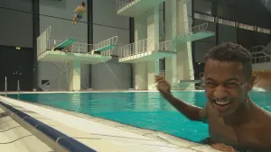 Foto van presentator die staat in een groot binnenzwembad met hoge duikplanken, van een lagere springt een man eraf.