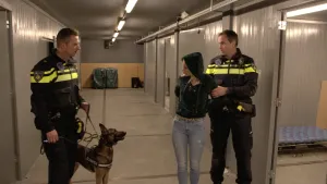 Foto van presentator in hondenkennel, twee politieagenten staan erbij, één ervan houdt een herdershond aan de lijn vast.