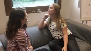 Foto van twee vrouwen die op een bank in een kantoorachtige ruimte zitten, ze praten met elkaar.