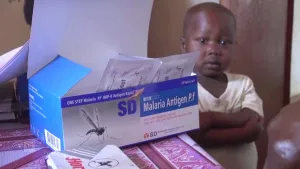 Foto van een zwart jongetje dat staat naast een tafel waarop een doos staat met middel tegen malaria.