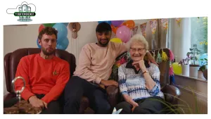 Foto van met ballonnen versierde huiskamer waar twee mannen zitten bij een vrolijke oude vrouw, oma.