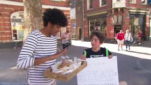 Foto van twee jongens die op straat bij een tafel staan waarop wat spullen liggen die ze te koop zijn, met papier erbij met "te koop" erop.