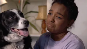 Foto van een huilend meisje naast een hond in de huiskamer, je ziet met name haar hoofd en van de hond zijn/haar kop.