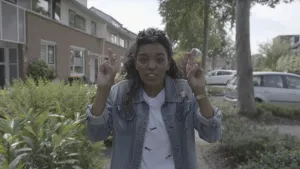 Foto van presentator Sosha die gebarentaal laat zien door met beide handen haar middelvinger te kruisen met de wijsvinger, op straat.