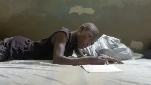Foto van een zwarte jongen die op zijn buik op bed ligt en een brief schrijft.