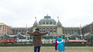 Foto van man en meisje die op het strand staan, je ziet ze van achteren, met op de achtergrond groot gebouw, hotel, het Kurhaus te Scheveningen waarnaar ze kijken.