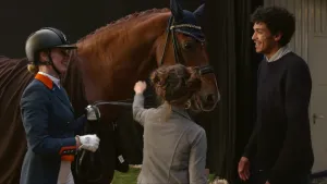 Foto van presentator, vrouw in paardrijkleding en een meisje die staan bij een groot bruin paard, het meisje borstelt het paard.
