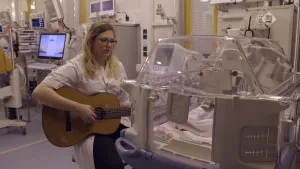 Foto van vrouw die gitaar speelt bij een couveuse met een baby erin, in een ziekenhuiskamer.