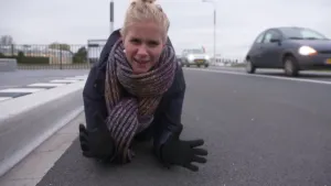 Foto van presentator die op haar buik op snelweg naast de vangrail ligt waar auto's op rijden.