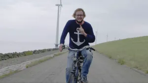 Foto van presentator op de fiets die rijdt langs een dijk met hoge windmolens erlangs.