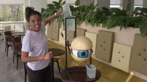 Foto van presentator in kamer die staat bij een groot model van een oog en giet er uit bakje water overheen.