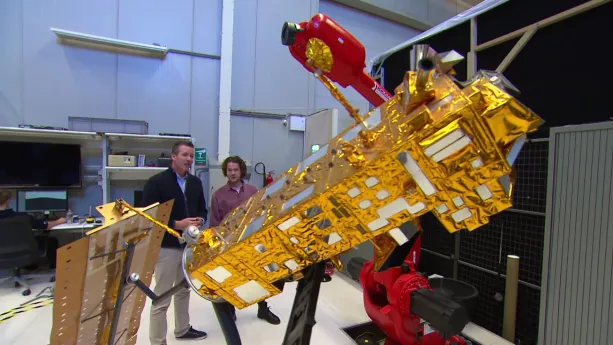 Foto van een oude satelliet in grote hal met twee mannen erbij.