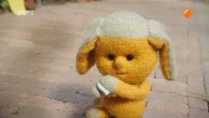 Foto van Dropje (pop) die buiten op de stoep/straat staat die een gehoorapparaatje vasthoudt.