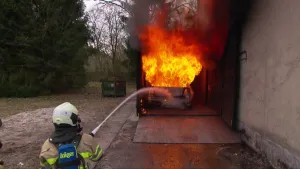 Foto van brandweerman met helm op die een in brand staande auto blust, het is een oefening, op oefenterrein.