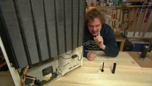 Foto van presentator die zit aan een tafel met een koelkast erop, je ziet de achterkant ervan.