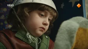 Foto, portret, van jongetje met een helm op, je ziet nog deel van het hoofd van Dropje (pop).