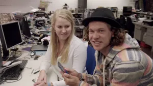 Foto van presentator met smartphone bij een vrouw die achter bureau zit, in een grote kantoorruimte.