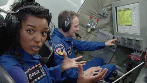 Foto van presentator die zit in simulator van een ruimtecapsule, naast haar zit een man met computerscherm die deze bedient.