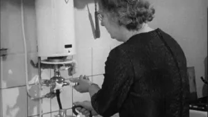 Oude foto, zwart/wit, van een keuken met een vrouw erin bij een ouderwetse geyser.