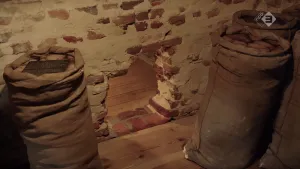 Foto van een bakstenen muur, houten vloer, met onderin een groot gat, daarachter zie je de houten vloer van ruimte achter die muur.