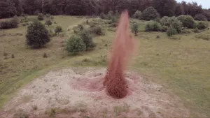 Foto van grasvlakte van bovenaf gezien waar in het zand een bom ontploft, veel zand spat omhoog.