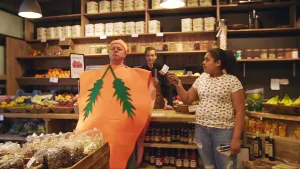 Foto van een winkel met toonbank, ervoor staat een man als een oranje wortel verkleed die wordt geïnterviewd door presentator.