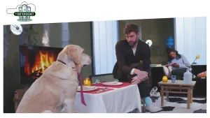 Foto van presentator die binnen aan gedekte eettafel zit waaraan ook een grote hond zit.