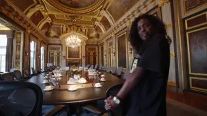 Foto van presentator die staat in grote zaal, de Treveszaal, met grote tafel erin met veel stoelen eraan.