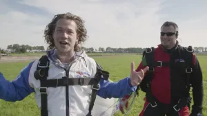 Foto van een presentator die net een parachutesprong heeft gemaakt staat in een wei en vertelt iets, naast hem staat een andere man met parachute met wie hij heeft gesprongen.