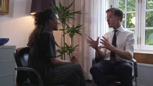 Foto van premier Mark Rutten die staat in kamer, van het Torentje, en praat tegen een vrouw die op een stoel zit.