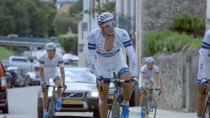Foto van drie wielrenners, hoofdzakelijk wit gekleed, die op een weg met huizen fietsen met erachter een auto die ze volgt.