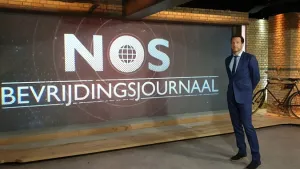 Foto van het NOS-Journaal met de nieuwslezer die in de studio staat met grote tekst op de muur "NOS Bevrijdingsjournaal".