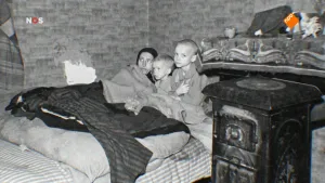 Foto van NOS-Journaal waarop oude zwart/wit foto van kinderen in een bed naast kachel in ouderwetse huiskamer.