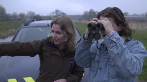 Foto van man en vrouw, de man kijkt door een verrekijker, bij een auto in de natuur.