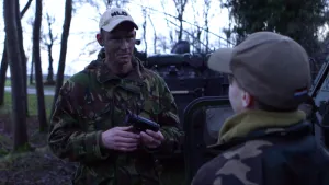 Foto van een soldaat met een verrekijker bij een legervoertuig in het bos, erbij staat een jongetje met een pet op.