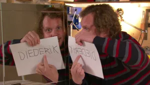 Foto van presentator die een vel papier met Diederik erop vasthoudt voor een spiegel waarin je het spiegelbeeld ervan ziet.