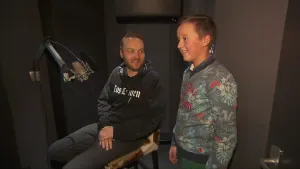 Foto van man die in een stoel zit met een microfoon ervoor, ernaast staat een meisje.