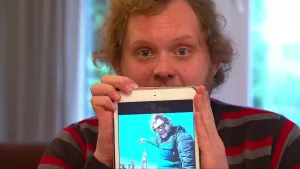 Foto van portret van presentator die een smartphone/mobiele telefoon vasthoudt met op het scherm een man buiten in Londen.