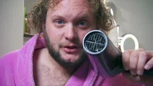 Foto van presentator, je ziet alleen zijn gezicht, die een föhn vasthoudt.