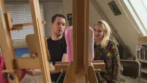 Foto van man en vrouw die achter een schildersezel staan waarop een klein schilderij staat.