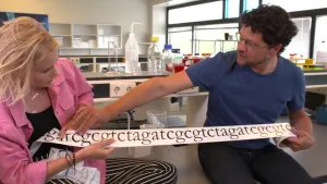 Foto van man en vrouw in een laboratorium die een reep papier vasthouden met erop reeks letters met de c, g, t en a.