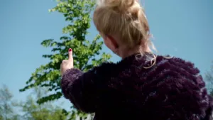 Foto van presentator van achteren gezien die met haar duim buiten naar een groene boom wijst.