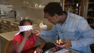 Foto van man aan tafel en geblinddoekt meisje dat iets gaat proeven wat die man haar aanreikt.