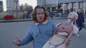 Foto van presentator die op buiten op straat een model van een mens, opengemaakt, waarbij je de organen ziet vasthoudt.