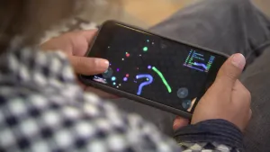 Foto van deel van een persoon, je ziet vooral zijn handen die een mobiele telefoon, smartphone, vasthouden waarop een spelletje.