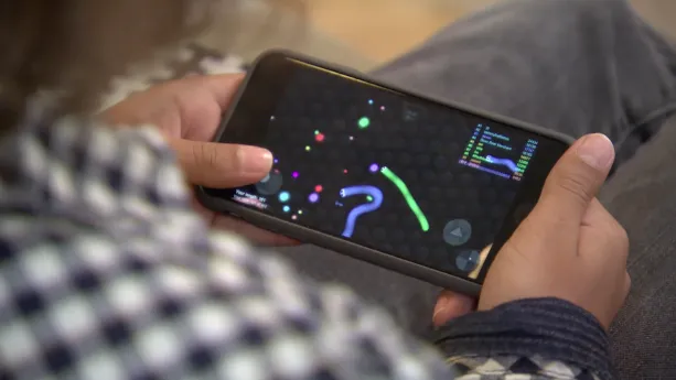 Foto van deel van een persoon, je ziet vooral zijn handen die een mobiele telefoon, smartphone, vasthouden waarop een spelletje.