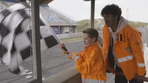 Foito van man en jongetje, met oranje werkkleding aan, die staan langs de baan van een racecircuit, het jongetje zwaait met een zwart/wit-geruite vlag.