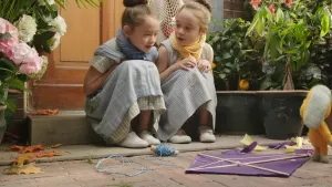 Twee meisjes die op de stoep zitten met voor ze een (kapotte) vlieger waarbij Dropje, een stoffen pop, staat.
